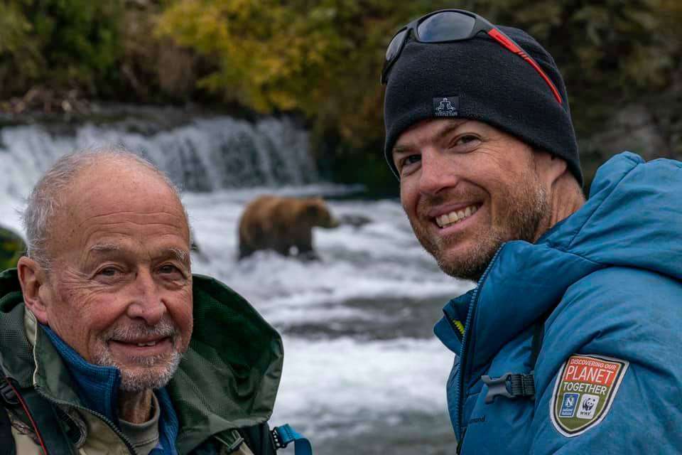 Guide and nature traveler pose with a bear in the background on Bears of Brooks Falls: A Photo Pro Expedition.