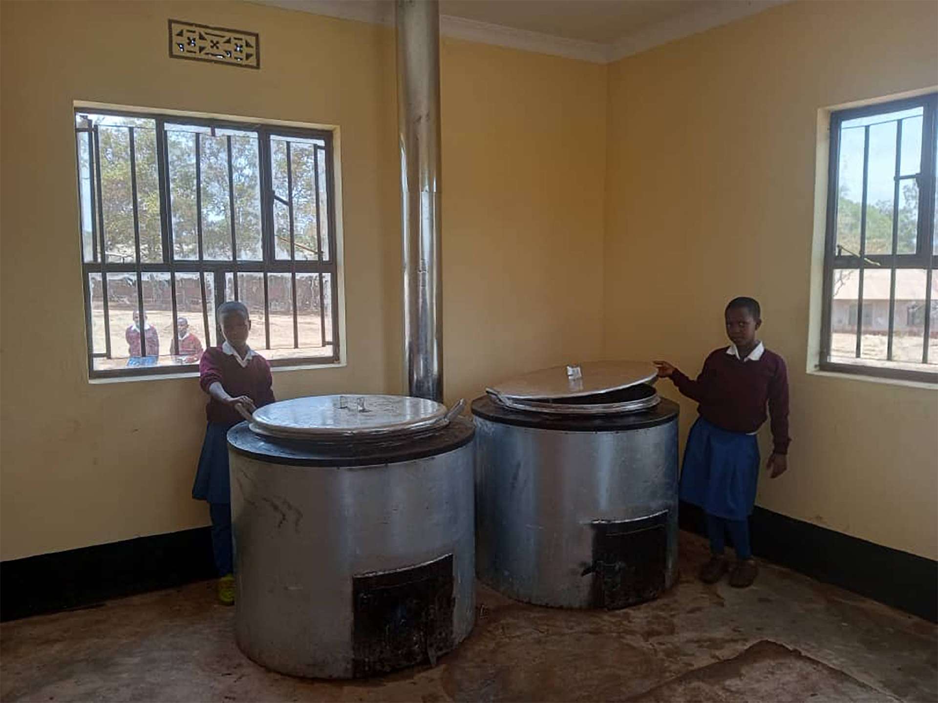 Two smiling children from a school in Tanzania take a photo next to donated kitchen materials from Nat Hab Philanthropy